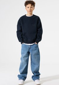 Sudadera azul marino con panel superior texturizado, corte holgado y puños acanalados, combinada con vaqueros anchos de color azul claro y zapatillas blancas.