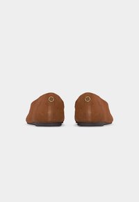Paire de chaussures plates en daim marron vues de dos avec de petits détails de logo doré rond sur les talons et des semelles noires.