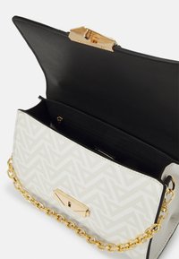 Sac blanc avec un motif géométrique, quincaillerie dorée et une bandoulière en chaîne. L'intérieur présente un revêtement noir avec une poche zippée.