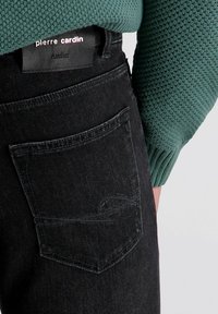 Schwarze Jeans aus Denim mit einer Gesäßtasche und gesticktem Detail, kombiniert mit einem strukturierten grünen Pullover. Schwarzes Lederlabel am Bund.