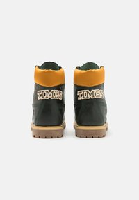 Tumedad roheline nahast saapad, millel on oranž pehme krae, beež väliskülg ja tikitud "TIMBS" logo seljal. Tekstuuriga viimistlus, kõrge disain.