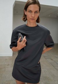 Temno srebrna oversize obleka s kratkimi rokavi in okroglo ovratnikom, ki se ponaša z gladko teksturo. Model drži clutch v eni roki.