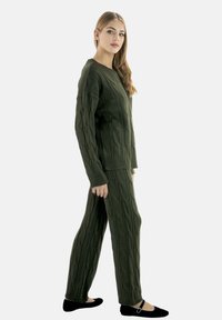 Grønt strikket sæt med sweater og bukser med kabelmønster; har lange ærmer, løs pasform og flade sorte sko med dekorativ rem.