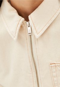 Off-white blusjacka med krage, tillverkad av texturerat tyg. Har en framzipper med ett silverfärgat drag och kontrasterande orange sömmar.