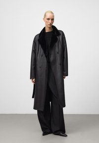 Zwart leren dubbelzijdig jasje met een zacht zwart nepbont kraag, oversized silhouette en lange mouwen. Draagt over bijpassende zwarte kleding.