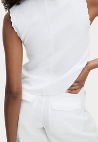Top bianco senza maniche con scollatura a zig-zag, realizzato in tessuto leggero, caratterizzato da un semplice design sul retro e discreti dettagli di cucitura.