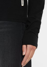 Schwarzes Langarmshirt aus weichem Stoff, mit enganliegendem Bündchen und einem seitlichen Etikett. Kombiniert mit dunkler Jeans.