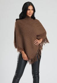 Brun stickad poncho med hög hals, med texturerat tyg och fransade kanter. Matchad med åtsittande svarta fusk läderbyxor.