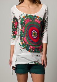 Vit långärmad skjorta med ett färgglatt cirkulärt mönster i rosa, grönt och blått; har en sidoknytning. Matchas med mörkgröna shorts.