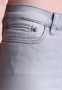 Un jean en denim gris clair présente une poche avant avec un subtil détail de couture, des boutons métalliques, et une texture lisse.