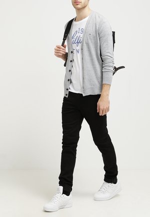 Mies pukeutuneena harmaaseen neuletakkiin valkoisen graafisen t-paidan päällä, mustiin housuihin, valkoisiin tennareihin ja mustaan reppuun, kävelee yksiväristä valkoista taustaa vasten.