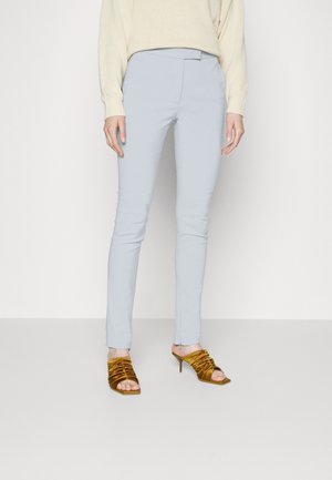 Osoba ubrana w jasnoniebieskie wąskie spodnie, kremowy sweter i musztardowe sandały na obcasie z marszczeniem, stojąca na tle gładkiej białej ściany.