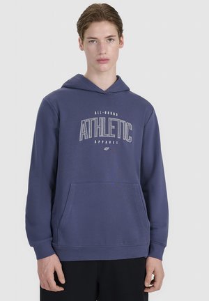 Młoda osoba ubrana w niebieską bluzę z kapturem z przednimi kieszeniami i tekstem "ATHLETIC" na piersi, stojąca na jednolitym tle.