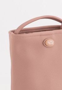 Rosa texturerad konstläderväska med en strukturerad form, utrustad med ett handtag överst och en guldfärgad cirkulär logotyputstickare på sidan.