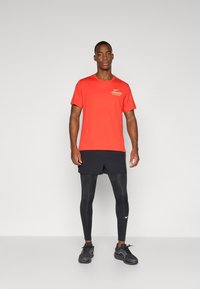 Κόκκινο αθλητικό μπλουζάκι Nike με το "LONDON" τυπωμένο σε κίτρινο, συνδυασμένο με μαύρες βερμούδες και μαύρα συμπιεστικά κολάν. Τα παπούτσια είναι σε μαύρο αθλητικό σχέδιο.