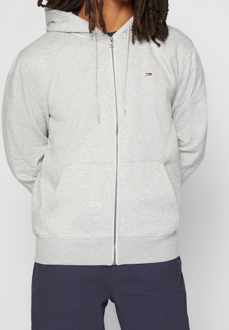 Hellgraue Zip-up Kapuzenjacke aus weichem Material, mit einer Fronttasche, gerippten Bündchen und einem kleinen Logo auf der Brust.