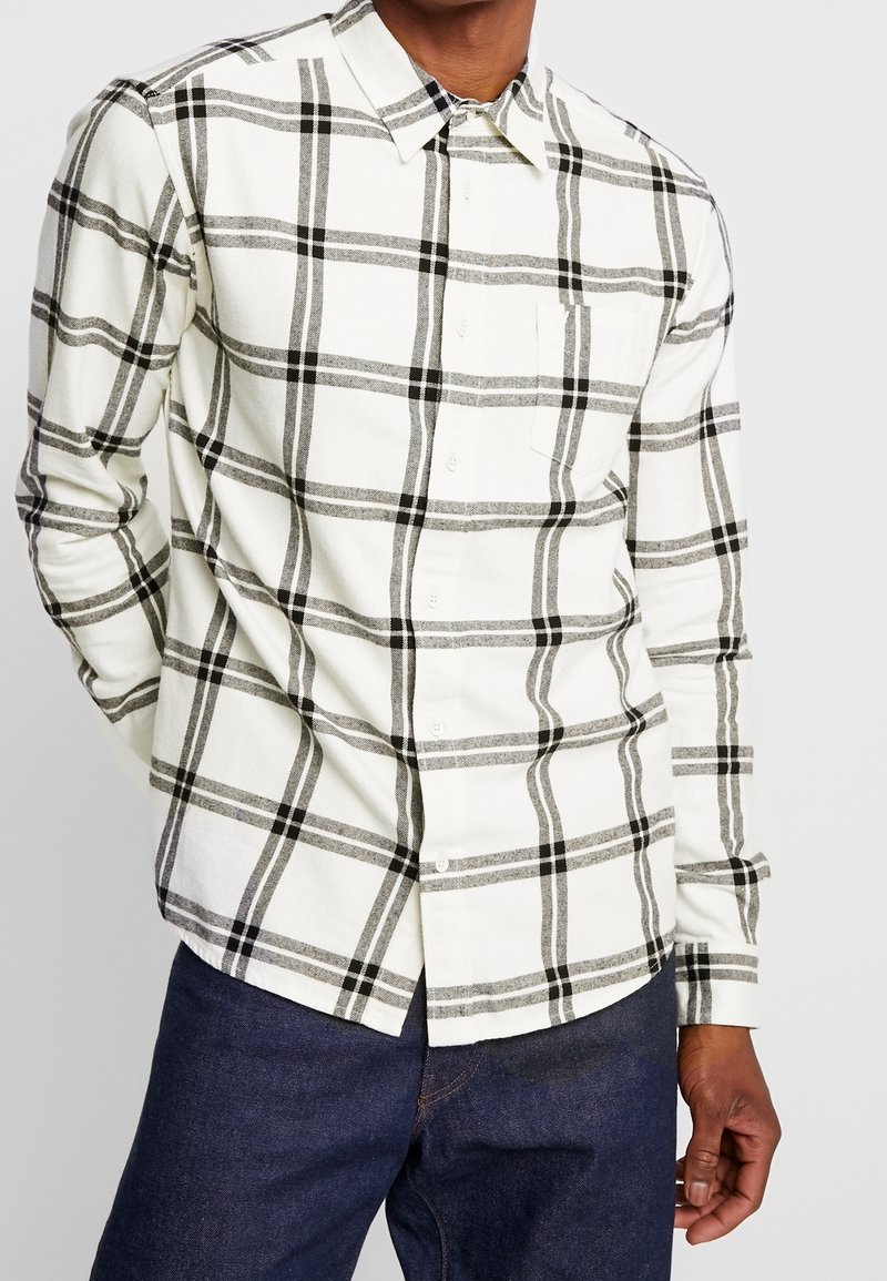 Homme portant une chemise blanche boutonnée avec un motif à carreaux noirs et gris et un jean bleu foncé, debout avec une main derrière le dos.