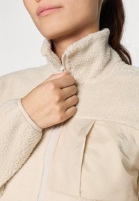 Chaqueta de forro polar beige con un alto cuello, media cremallera y panel de tela suave, que cuenta con un bolsillo de parche y un sutil detalle de logo.