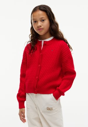 Young girl with long hair wearing a red knit cardigan and white pants with Tommy Hilfiger embroidery, standing with one hand in pocket.