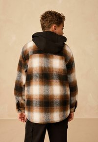 Checked flannel shirt in brown, black, and white with a black hood. Features button cuffs and a rounded hem. Soft texture.