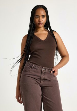 Ung kvinde med langt flettet hår iført en mørkebrun ærmeløs top og højtaljede brune jeans stående foran en ensfarvet baggrund.
