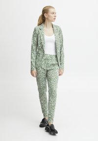 Groene jurk met een wit stippenpatroon, bestaande uit een op maat gemaakte blazer en slim-fit broek. Draagt met een wit top en zwarte instappers.