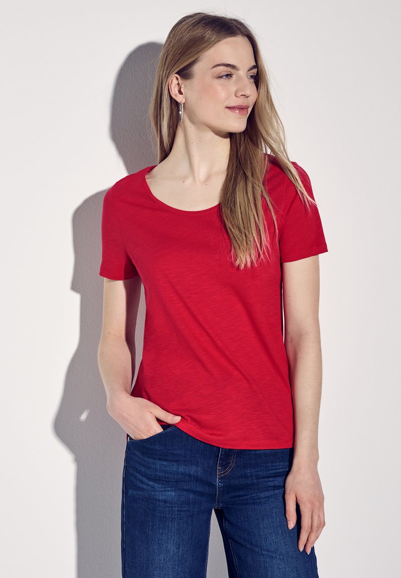 Rød T-shirt med korte ærmer og rund hals, lavet af et blødt, tekstureret stof. Kombineret med blå jeans, hvilket fremhæver et afslappet look.