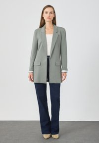 Blazer vert clair avec deux poches avant, épaulettes structurées et manches retroussées, associé à un jean large bleu foncé.
