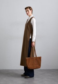 Braunes Wollkleid mit seitlichen Schlitzen, getragen über einem weißen Hemd. Kombiniert mit dunkelblauen Hosen und einer braunen Ledertasche. Silberschuhe sind sichtbar.