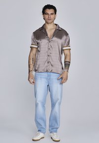 Camisa de satén de manga corta, de color marrón claro, con un patrón sutil y puños a rayas doradas, combinada con pantalones vaqueros de mezclilla azul claro y zapatillas blancas.