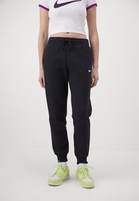 Pantalones de chándal negros con cintura con cordón y bolsillos laterales, combinados con una camiseta blanca corta que presenta un acento púrpura y el logo de Nike.