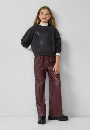Mädchen mit langen blonden Haaren, das einen dunklen Pullover mit Schriftzug, bordeauxfarbene Kunstlederhosen und schwarze Stiefel trägt und mit einer Hand auf der Hüfte steht.