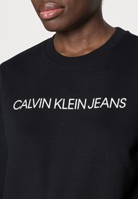 Černá mikina s vroubkovaným crew krkem, vyrobená z texturované tkaniny, s potiskem "CALVIN KLEIN JEANS" tučnými bílými písmeny.