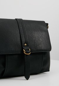 Borsa a tracolla in pelle nera con pattina ribaltabile, dettagli in metallo dorato, superficie testurizzata, tracolla regolabile e base cilindrica.