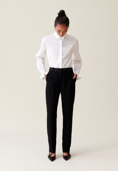 Camicia bianca a maniche lunghe con colletto arricciato, abbinata a pantaloni neri su misura. Scarpa nera a punta completano l'outfit.