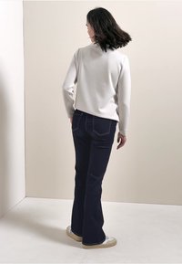 Pull gris clair, jean évasé bleu foncé, baskets blanches. Tissu lisse, coupe décontractée, poches arrière standard avec surpiqûres contrastées.