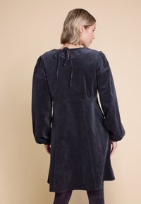 Mørk grå corduroy kjole med lange pufferm og en lav rygg med knyting. Har en utformet design og teksturert stoff.