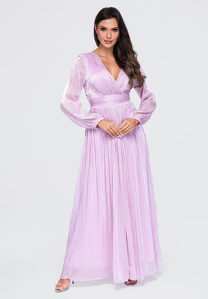 Mujer con cabello largo y oscuro que lleva un vestido largo plisado en color lavanda brillante, con mangas largas y escote profundo en V, de pie con las manos entrelazadas.