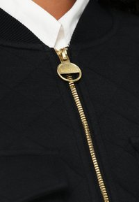 Μαύρο καπιτονέ μπουφάν με χρυστό φερμουάρ και στρογγυλή τσοκέν, με λευκό πουκάμισο με γιακά ορατό από κάτω.