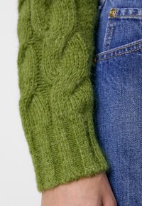 Pull en maille verte avec un motif de câbles texturé et des poignets côtelés, associé à un jean en denim bleu. Gros plan sur les détails de la manche.