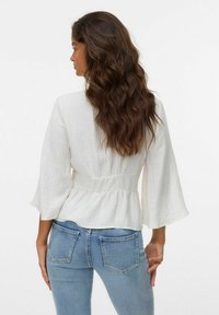 Donna con lunghi capelli castani mossi, che indossa una blusa bianca a maniche larghe e jeans attillati di colore blu chiaro, rivolta di lato contro uno sfondo semplice.