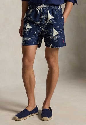 Tmavomodré šortky s bielymi vzormi plachetníc, šnúrka v páse, nosené s tmavomodrými espadrilkami a čiastočne viditeľnou tmavomodrou košeľou.