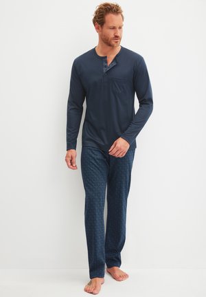 Man in navy long-sleeve henley shirt and patterned navy pajama pants standing barefoot on a white floor against a white wall.