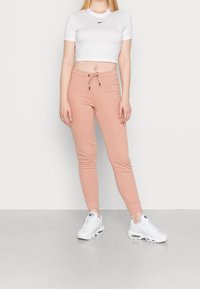 Kvinna klädd i en vit kortärmad Nike-t-shirt, persikofärgade joggers med dragsko och vita Nike Air Max-sneakers som står på ett grått golv.