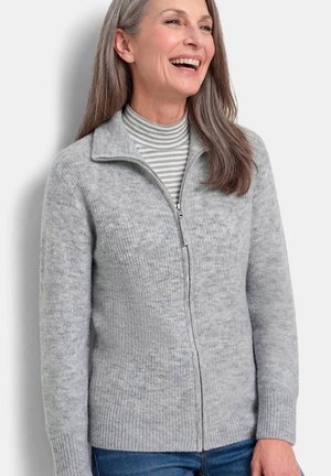 Frau lächelt, trägt einen hellgrauen Zip-Cardigan über einem gestreiften Rollkragenpullover, kombiniert mit blauen Jeans, steht vor einem einfarbigen Hintergrund.
