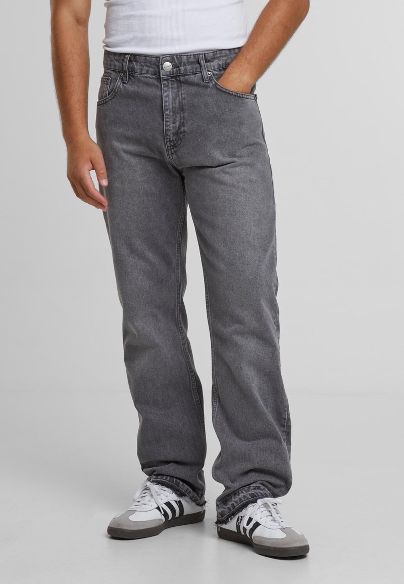 Jean en denim gris à coupe décontractée, cinq poches, fermeture par bouton et détails de coutures visibles, porté avec des chaussures de sport blanches et grises.