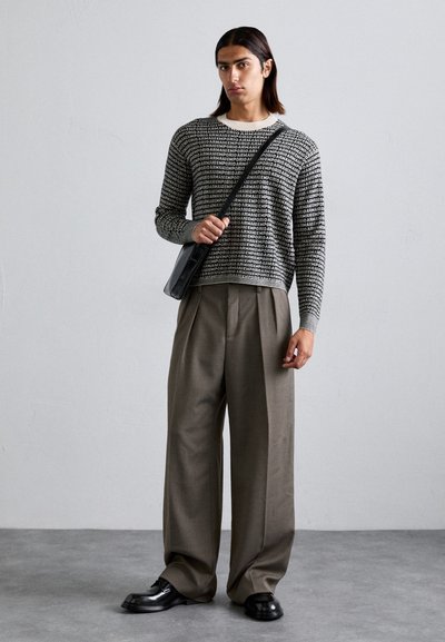 Pulover tricotat cu un model de text alb-negru, asortat cu pantaloni largi bej. Ține o geantă de umăr neagră și poartă pantofi negri lucioși.