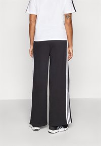 Pantaloni de trening lungi, negri, cu dungi albe pe laterale, asociați cu un tricou alb cu mânecă scurtă. Pantofii sport sunt vizibili în partea de jos.