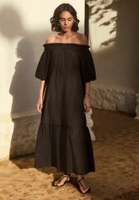 Zwarte maxi-jurk met off-shoulder, pofmouwen, gelaagd ontwerp en textuurstof. Draagt in combinatie met zwarte sandalen met decoratieve bandjes.