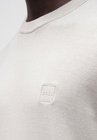 Detailný záber na osobu nosiacu svetlobéžový pletený sveter s malou štvorcovou nášivkou s logom "BOSS" na hrudi.